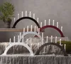 A collection of arched candle holders with electric candles, arranged on a draped surface with a plant in the background. Produktbild-Vorschau