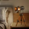 A vintage-style spotlight lamp on a tripod stand sits on a bedside table next to a bed. Náhled obrázku produktu
