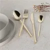 A set of champagne-colored cutlery arranged on a textured gray surface, with a white bowl and saucer in the background and dried flowers in the upper right corner. Anteprima dell’immagine prodotto