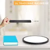 A hand holding a thin black object, with a book and a round light fixture on a table below. The background is a blurred interior scene. Voir les images du produit