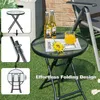 A round glass-top bistro table with a black metal frame, set up on artificial grass, with two bottles of beer, a glass of juice, and a sunflower on top. The table is shown in use in an outdoor setting. Náhľad obrázku produktu