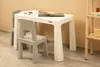 A child's table and two chairs are set up in a room with a light beige wall and a wooden shelf unit to the right. Náhled obrázku produktu