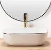 A white oval sink with a gold rim and a gold faucet is shown in a bathroom setting with a round mirror in the background. Náhľad obrázku produktu