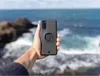 A hand holds a phone case with a mount in front of a blurred ocean background. Produktbild-Vorschau
