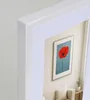 A framed picture of a red poppy on a blue background, displayed in a room with a sofa and a small plant. Produktbild-Vorschau