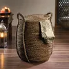 A woven basket with a lid and a folded cloth draped over it, placed on a wooden floor next to a lantern and a table with candles. Produktbild-Vorschau