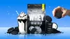 A bag of protein powder is shown next to a milkshake and several cookies, with a hand holding a cookie in the background. Produktbild-Vorschau