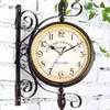 A double-sided antique-style station clock mounted on a white brick wall with green leaves in the background. Produktbild-Vorschau