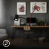 A black floor lamp with a flexible head is shown in a modern office or living room setting. The lamp head is depicted in two positions, indicating its adjustability. A circular icon with arrows is present in the bottom left corner. Anteprima dell’immagine prodotto