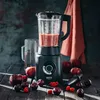 A blender filled with a pink smoothie, surrounded by apples, plums, and raspberries on a wooden table. Produktbild-Vorschau