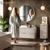 A woman in a bathrobe stands in a modern bathroom, looking into a round mirror above a vanity. The bathroom features a bathtub, a vanity with drawers, a tall cabinet, and decorative elements. Produktbild-Vorschau