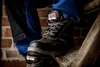 A close-up shot of a person wearing black work boots and blue work pants, with the boot resting on a wooden beam. Produktbild-Vorschau