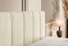 A close-up view of a beige upholstered headboard of a bed, with a bedside lamp and decorative reeds in the background. Produktbild-Vorschau