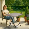 A woman sits on a chair next to a folding table on a patio, with a garden in the background. Produktbild-Vorschau