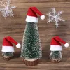 Three small, artificial Christmas trees decorated with Santa hats, placed on a wooden surface with decorative snowflakes in the background. Náhled obrázku produktu