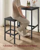 A person is sitting on a bar stool at a bar table, with bottles and a vase on the table. Produktbild-Vorschau