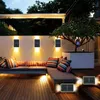 An outdoor patio area at dusk, illuminated by solar wall lights and accent lighting under a sofa. The scene features a sectional sofa with colorful cushions, a wooden deck, and a potted tree. Náhľad obrázku produktu