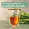 A glass of amber liquid, likely salmon oil, is presented in front of a blurred background of green grass-like elements and a light-colored wall. Produktbild-Vorschau