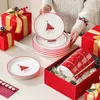 Eine Person packt das Geschirrset mit Weihnachtsmotiven in rot-goldene Geschenkboxen am Tisch ein. Voir les images du produit