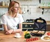 A woman in a kitchen looking away from the camera, with a sandwich maker and ingredients on the counter. Náhľad obrázku produktu