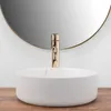 A white vessel sink with a gold faucet in front of a round mirror. Produktbild-Vorschau