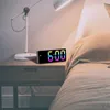 A digital alarm clock with a colorful display sits on a wooden nightstand next to a lamp and a bed. Náhled obrázku produktu
