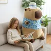 A young girl is sitting on a couch reading a book next to a large capybara plush toy. Produktbild-Vorschau