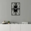 A black metal wall clock with Roman numerals is mounted on a light gray wall above a white cabinet. The cabinet has a vase, a small potted plant, and books on top. Produktbild-Vorschau