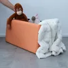 A person's hand is placing a stuffed orangutan into an orange storage bench. A white blanket is draped over the side of the bench. Anteprima dell’immagine prodotto