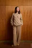 A woman stands in front of a wooden wall, wearing a beige knitted sweater and matching pants. Produktbild-Vorschau