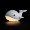 A white whale-shaped night light with a starry pattern on its back, emitting a warm yellow glow from its belly, set against a black background. Produktbild-Vorschau