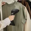 A person is using a handheld garment steamer on a green t-shirt hanging on a rack. Podgląd zdjęcia produktu