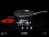 A frying pan is placed on a stovetop with a gas burner lit, and electric heating elements glowing red. The background is black. Náhled obrázku produktu