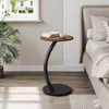A small round side table with a curved black metal base and a wood-look top is placed next to a bed. A book and a cup of coffee are on the table. A potted plant is to the left. Produktbild-Vorschau