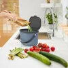 A person is shown discarding vegetable scraps into a blue compost bin in a kitchen setting, with fresh vegetables on the counter. Náhľad obrázku produktu