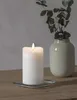 A white LED candle with a flame sits on a glass coaster on a wooden table, next to a vase with eucalyptus branches. Podgląd zdjęcia produktu