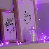 A shelf with two framed art prints and a candle, decorated with purple fairy lights. Produktbild-Vorschau