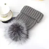 Graue Strickmütze mit großem schwarz-weißem Kunstfell-Pompon, neben einer weißen Teetasse