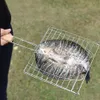 A person is holding a metal grill basket with a fish inside, outdoors on grass. Náhled obrázku produktu