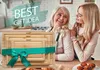 Two women in a kitchen, one older and one younger, smiling at each other. A set of wooden cutting boards is in the foreground, tied with a green ribbon. Produktbild-Vorschau