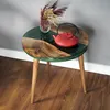 A round coffee table with a wooden and green epoxy top, featuring a red teapot and a vase of dried flowers. Produktbild-Vorschau