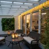 An outdoor patio area at dusk, featuring a table and chairs, a vertical garden, and string lights. Produktbild-Vorschau