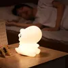 A person sleeping in bed with a dinosaur-shaped night light on a bedside table. Voir les images du produit
