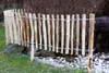 A rustic wooden garden fence made of vertical stakes bound together with wire, set in a garden with grass, rocks, and bushes. Produktbild-Vorschau