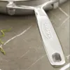 A close-up shot of a stainless steel pan handle with the brand name embossed on it, resting on a marble-like surface. Voir les images du produit