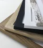 A close-up, angled view of a stack of four picture frames with different finishes: wood grain, black, white, and a distressed shabby white. Produktbild-Vorschau