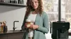 A woman is holding a coffee cup in a kitchen, with a coffee machine in the background. Náhled obrázku produktu