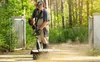 A man is using a gasoline-powered sweeper to clean a dirt path in a wooded area. Produktbild-Vorschau