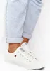 A person wearing white low-top sneakers and light blue cuffed jeans, with an anklet. Podgląd zdjęcia produktu