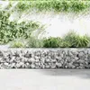 A gabion wall filled with stones, with plants growing on top and a white wall behind it. Náhľad obrázku produktu